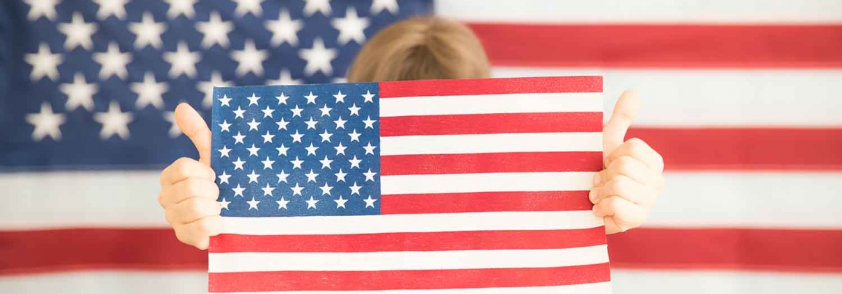 Young girl holding an American flag in front of an American flag backdrop.