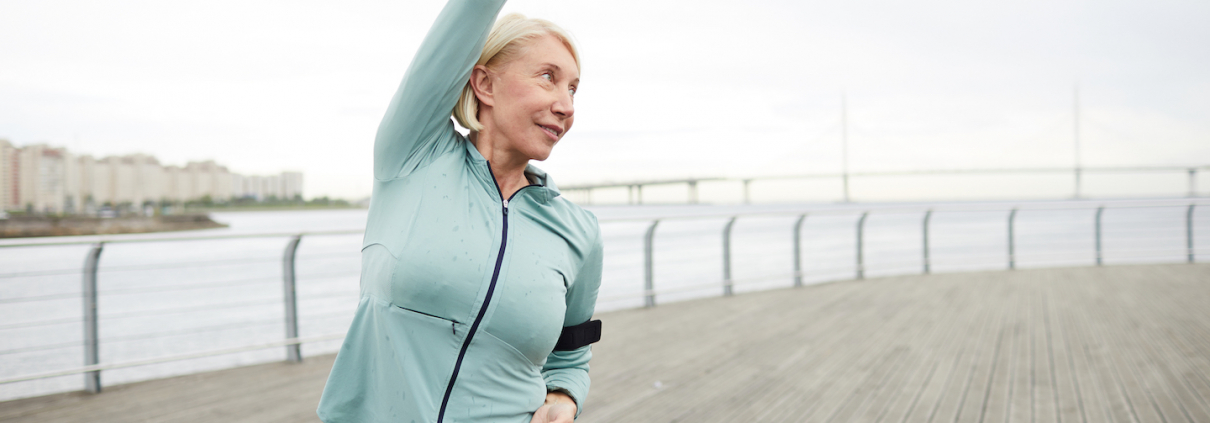 Mature active female in sportswear doing side-bends while training by riverside in urban environment on summer morning