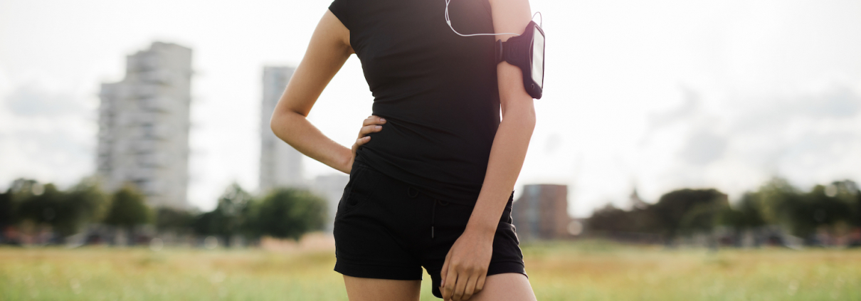 Cropped shot of fitness woman standing in urban park wearing sportswear and mobile phone armband. Female model in sportswear with earphones listening to music during workout break.