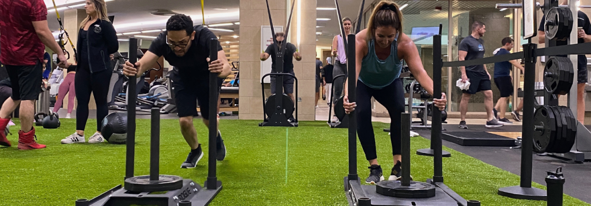 Two people on a turf pushing weighted sleds during a Faction workout class.