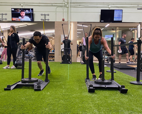 Two people on a turf pushing weighted sleds during a Faction workout class.