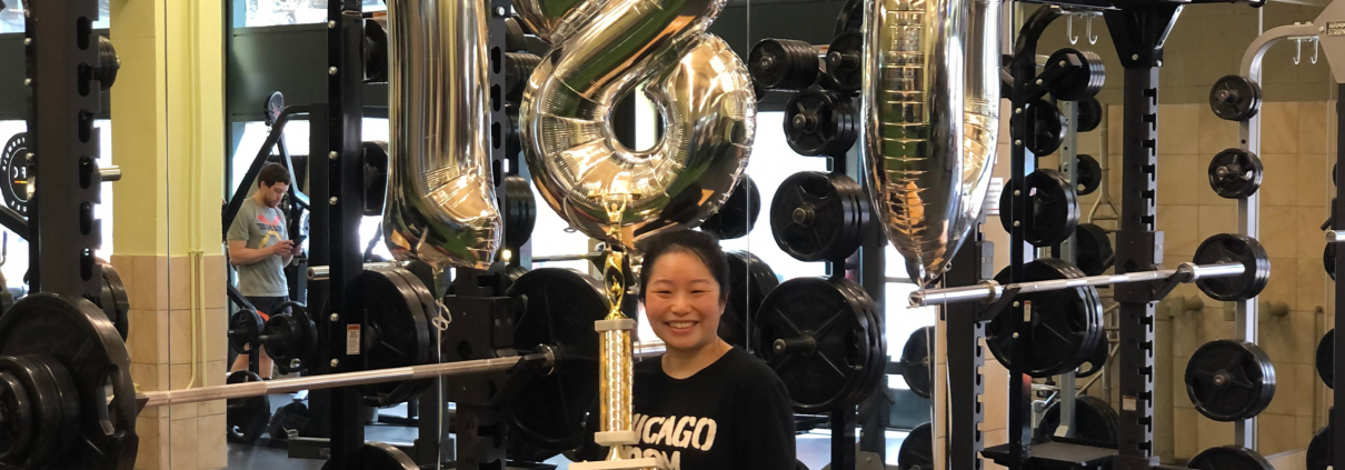 Emily With Balloons and A Trophy standing in front of a squat rack